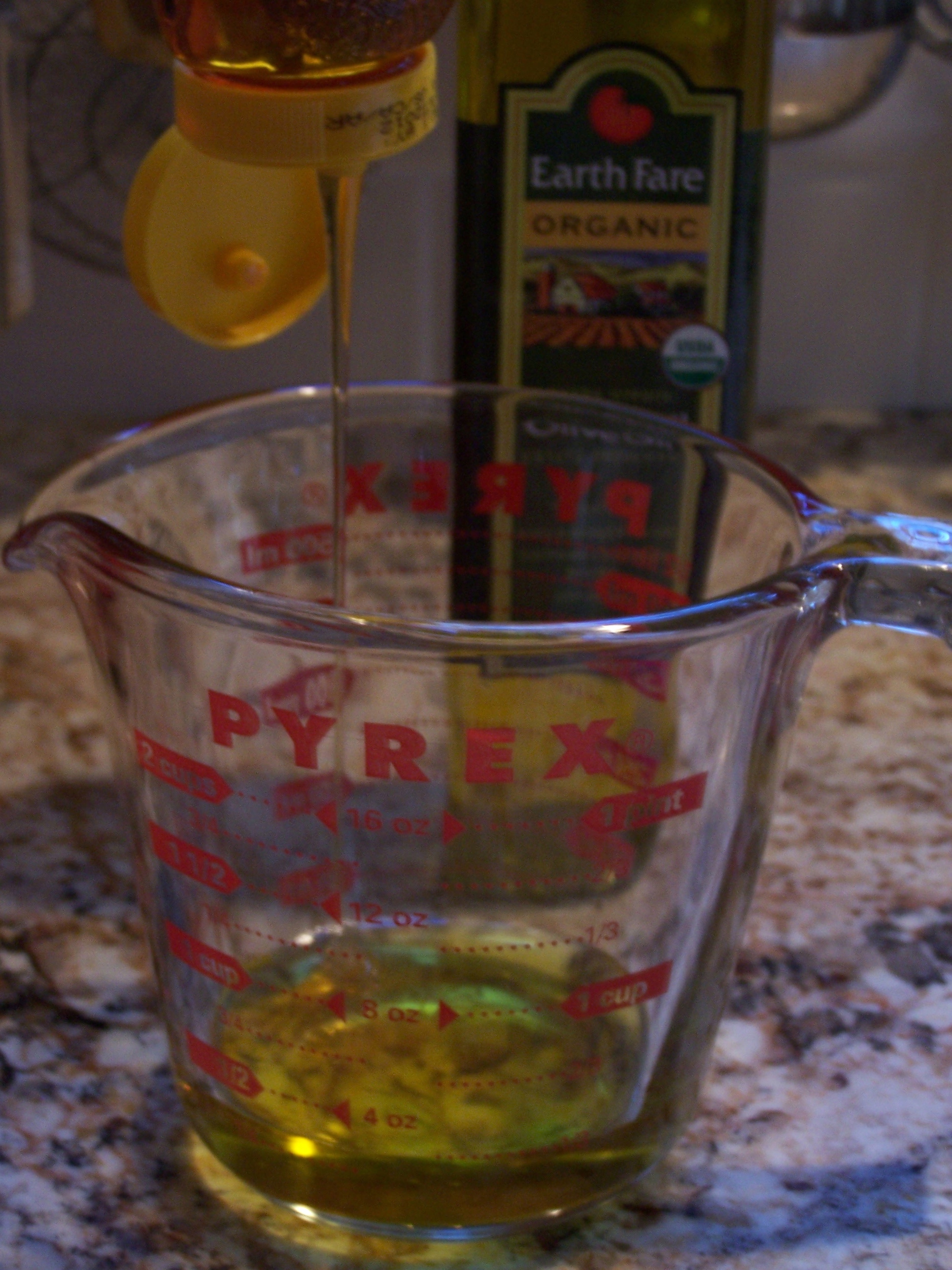 Measuring cup with honey being poured into it, olive oil on the counter