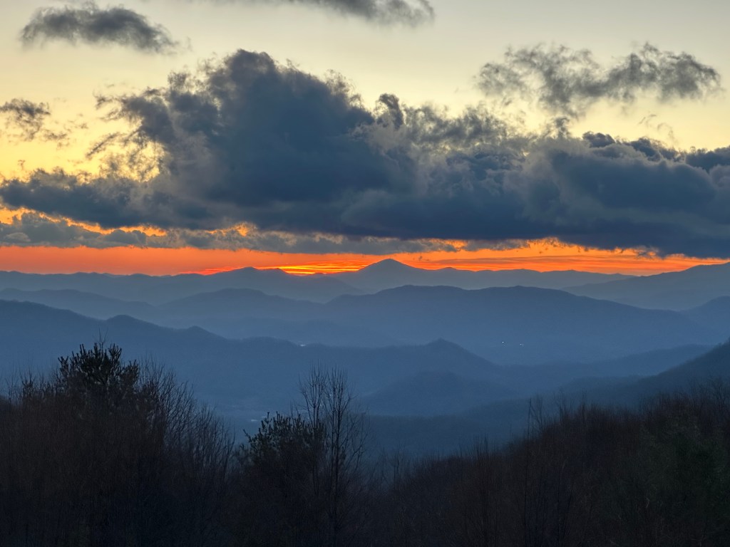 Mountains with sunrise in the distance, early morning fog, treetops, and swirling dark blue clouds