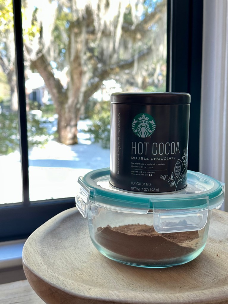 Photo of hot chocolate mix (homemade) in glass container, tin of starbucks hot cocoa mix on top of container, view out the window, snowy day in charleston, sc