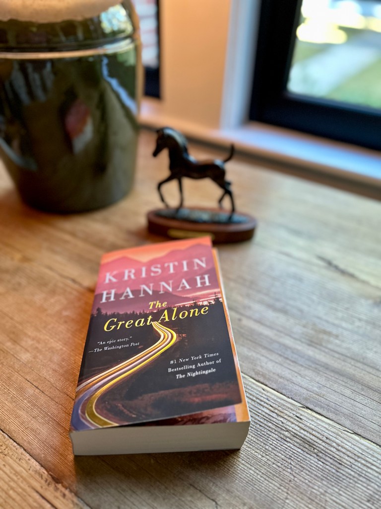 Book on table near window, The Great Alone by Kristin Hannah, cover of book shows mountains and a winding road, horse sculpture and lamp in background
