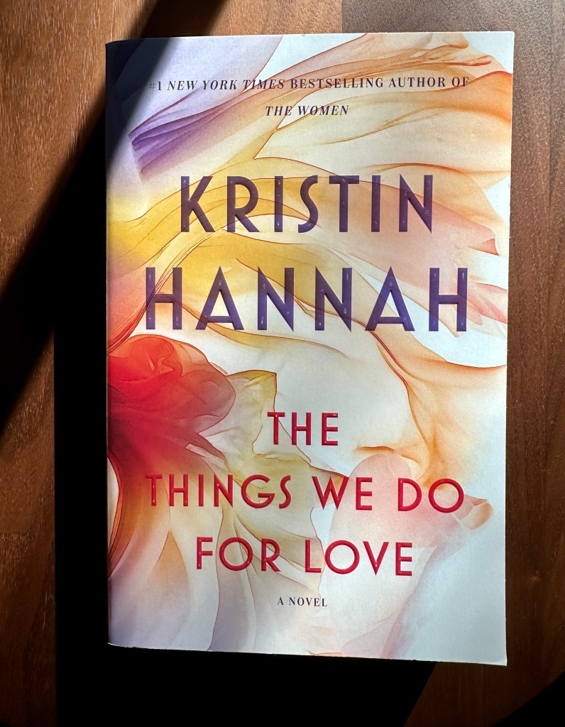 Paperback book on dark wood table, Kristin Hannah author, The Things We Do For Love is the title