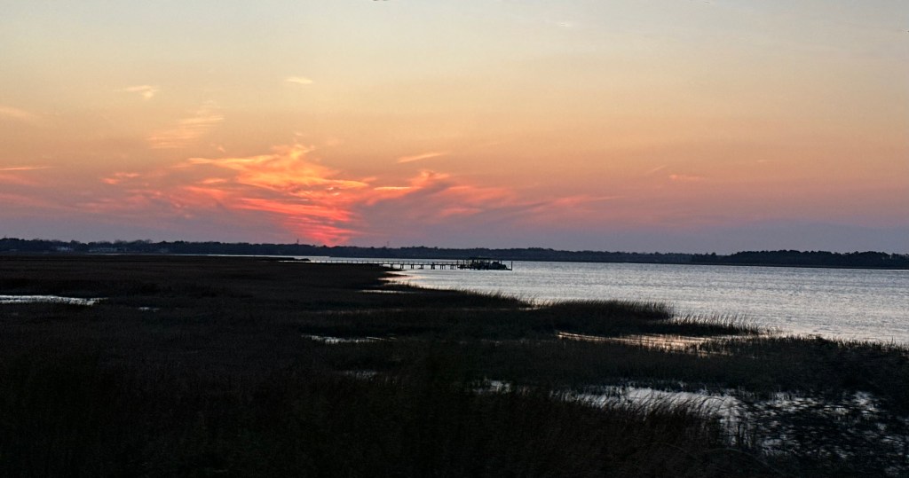 Marsh view at sunset, pink and orange skies, hints of purple along horizon, dock in the distance
