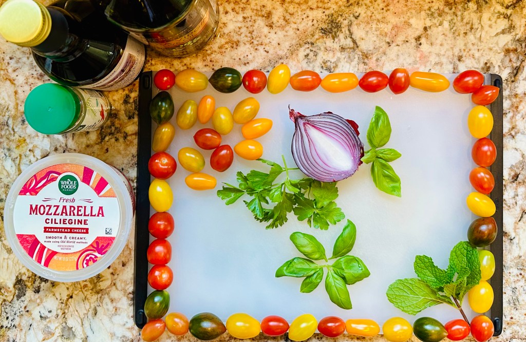 Cutting board, colorful grape tomatoes, red onion half, basil, parsley, mint leaves, mozzarella, pepper, balsamic vinegar, olive oil