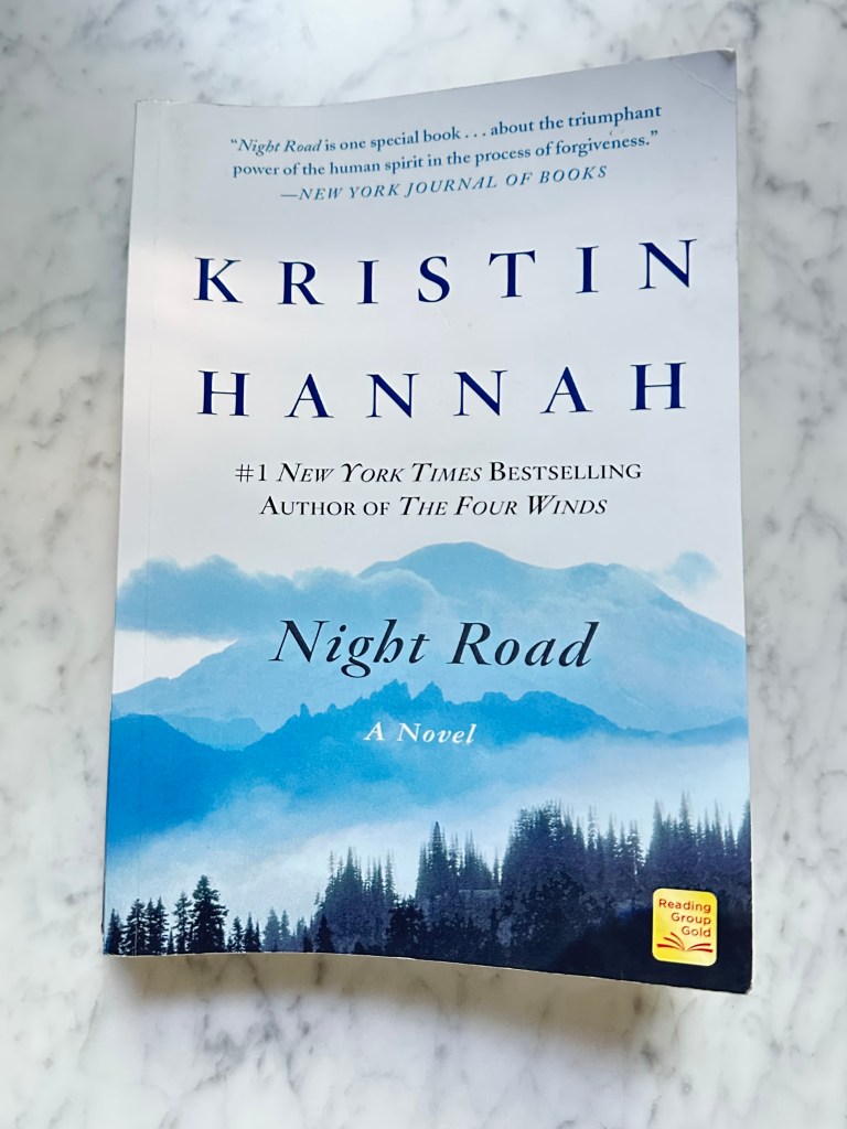 Book on marble table, Kristin Hannah Author, Night Road - A Novel, white cover with mountains, trees and clouds