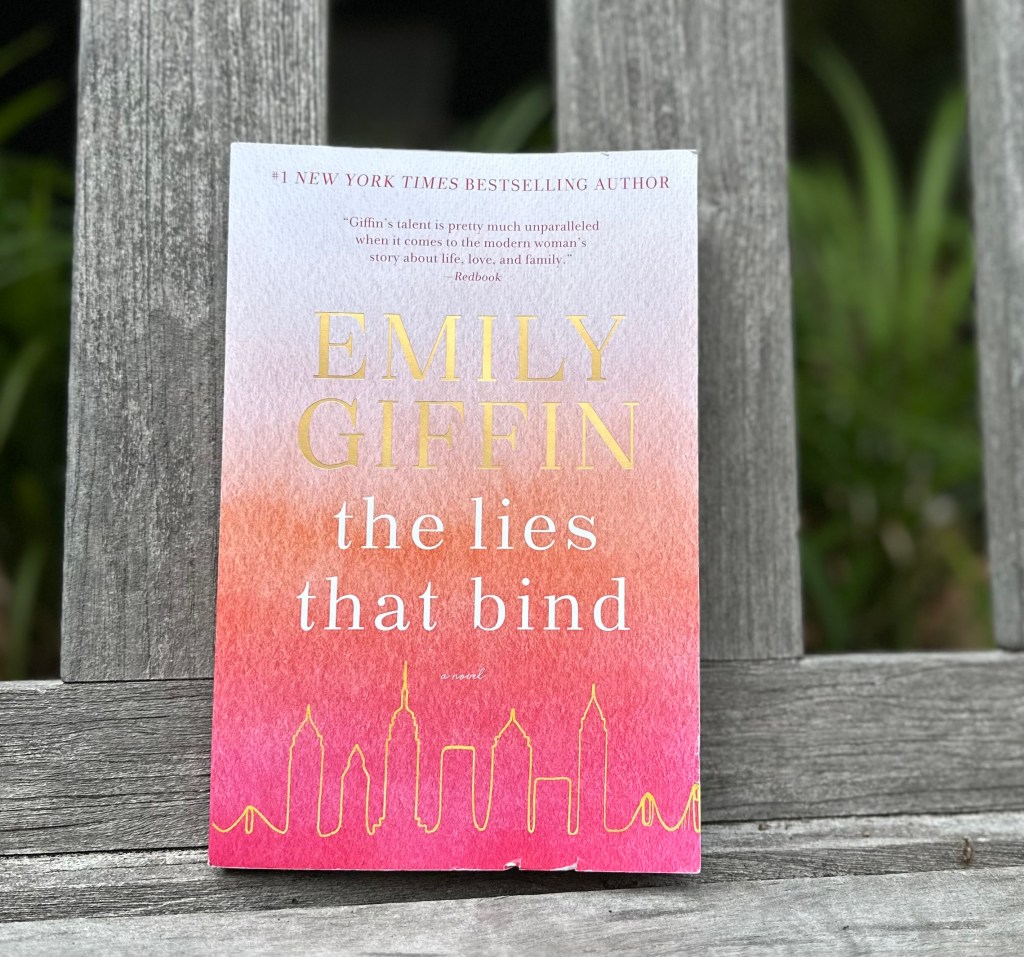 Book propped against teak sofa, liriope plant growing in the background, The Lies That Bind by Emily Giffin