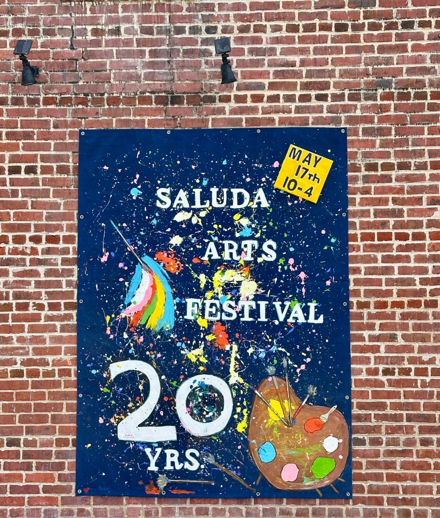 Blue sign on brick wall, reads Saluda Arts Festival 20 yrs, May 17th 10-4, colorful