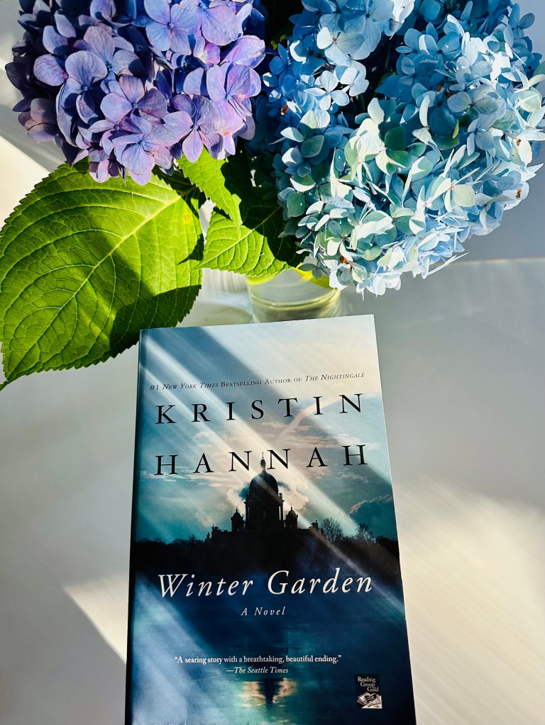 Book and hydrangea with purple flowers and green leaves on white table, Kristin Hannah, Winter Garden, A Novel