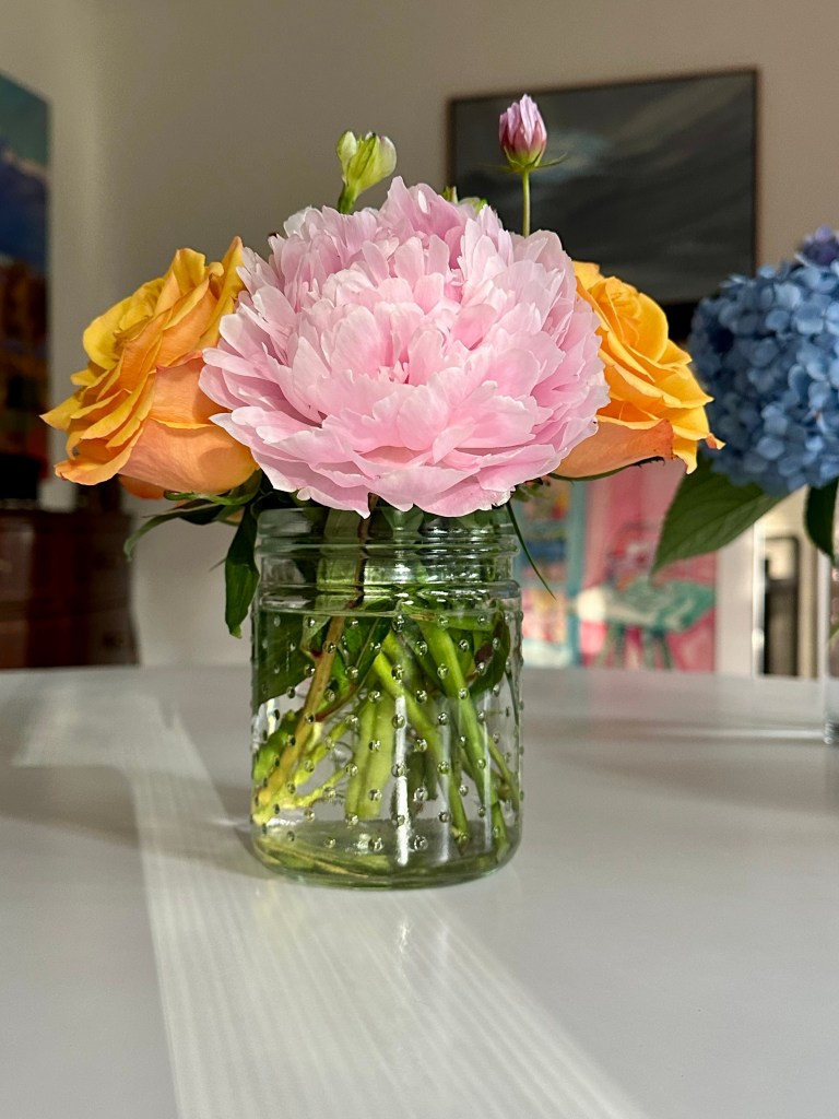 White table with vase of flowers, pink peony, two yellow roses, small pink daisy, pretty vase, paintings in the background 