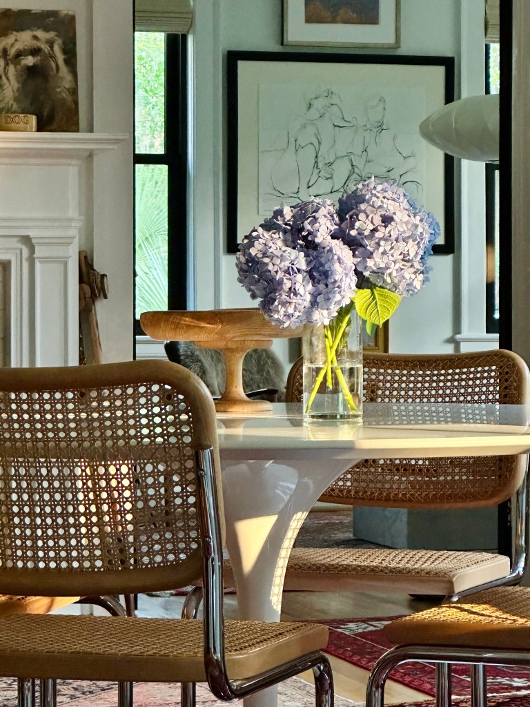 White table, cane chairs, a wooden nibble board, and a glass vase of lavender hydrangeas in water. 
