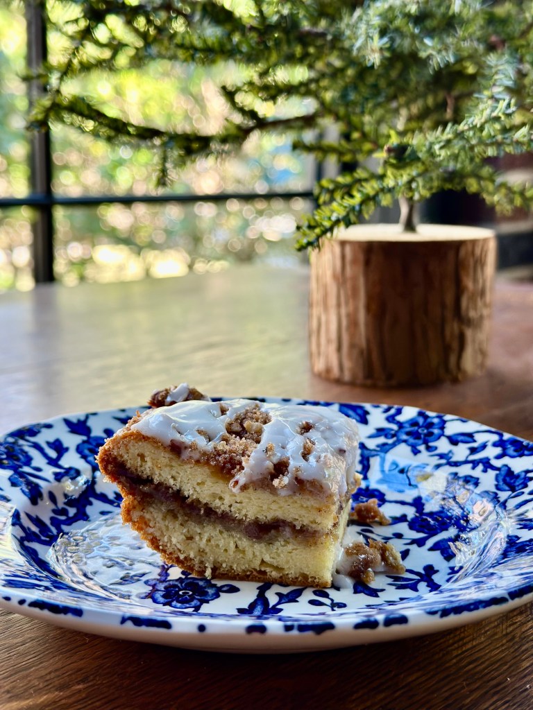 Coffee cake on blue and white plate in front of small tree near window.