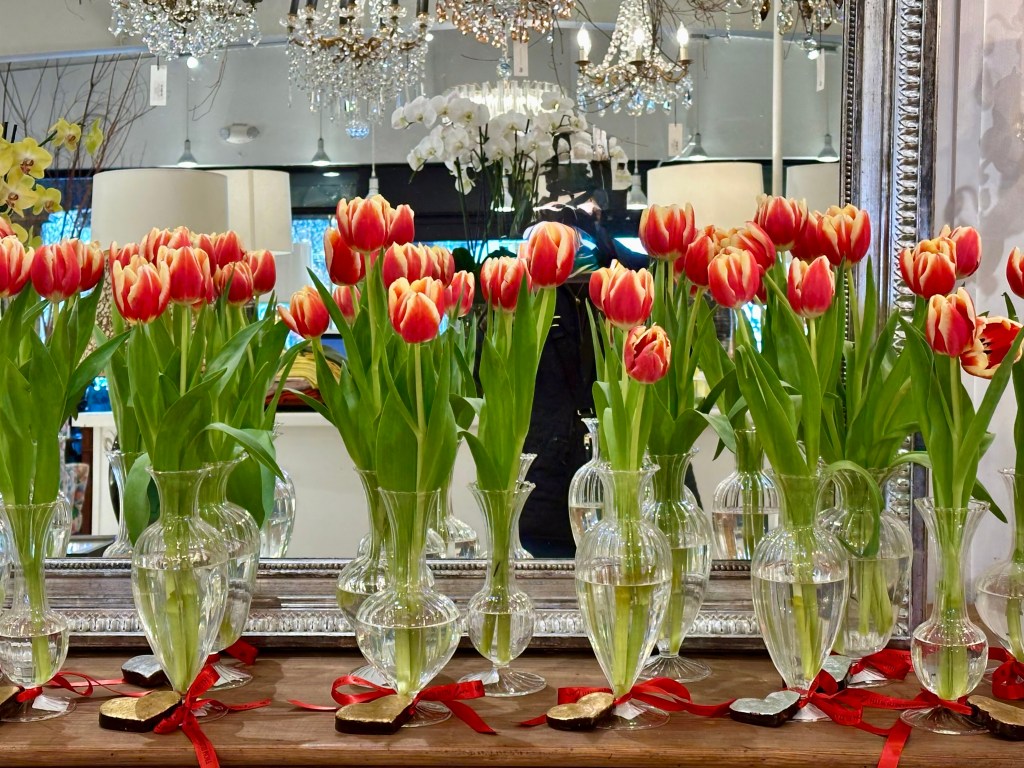 Vases lined up with tulips and a heart ornament in front of a mirror with a chandelier.